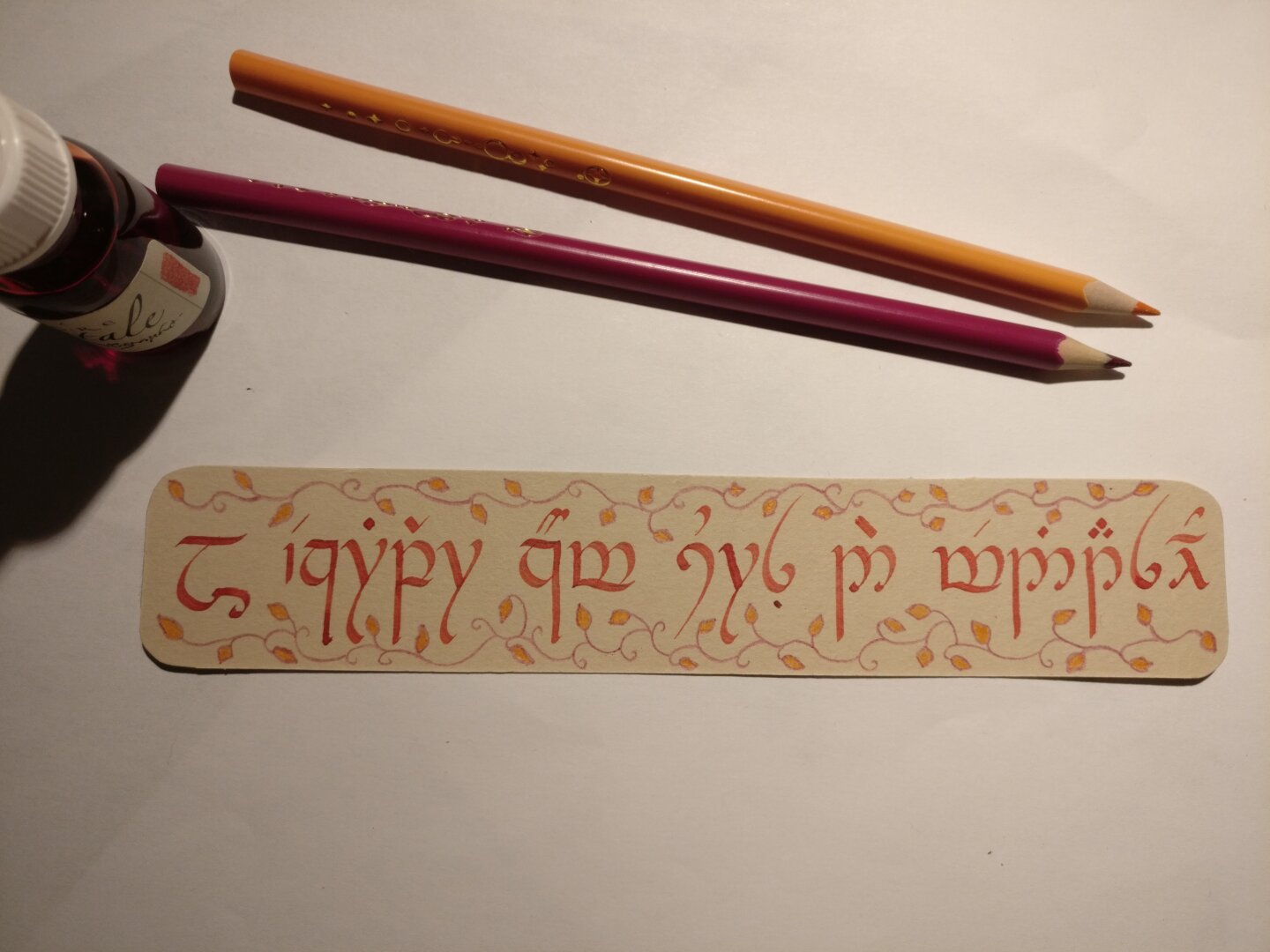 Un marque page avec du texte calligraphié en écriture Tengwar, posé à coté de deux crayons et d’un flacon d’encre rouge.